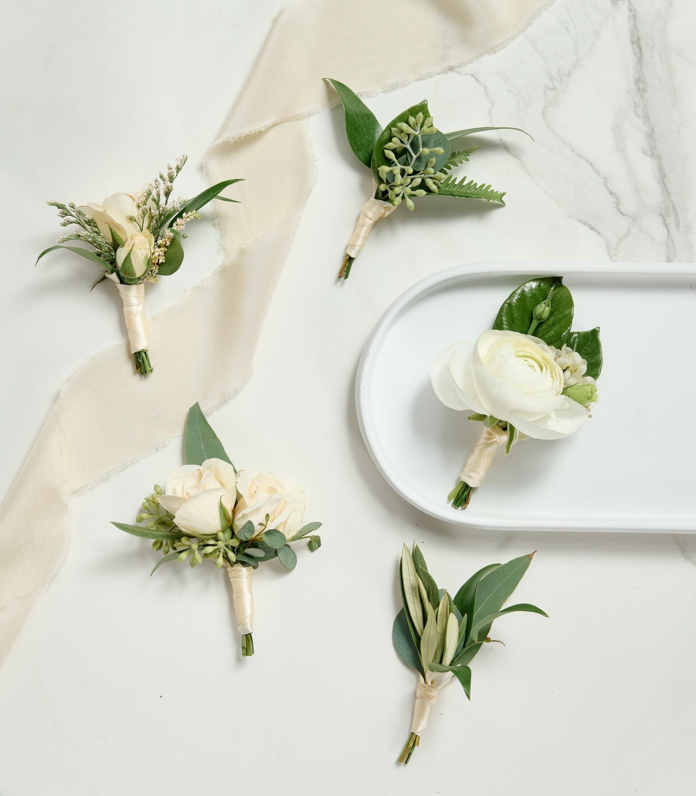 A collection of elegant white and cream boutonnieres with greenery, wrapped in ivory ribbon, displayed on a marble surface with a soft fabric accent.