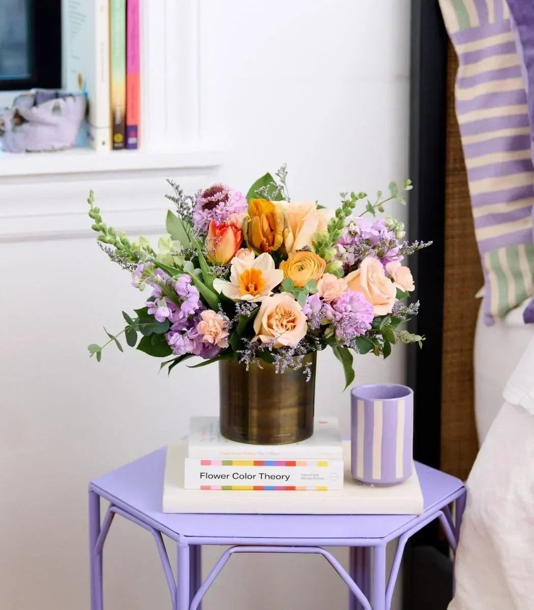 Pastel floral arrangement in a bronze vase displayed on a purple nightstand beside a bedroom lamp.