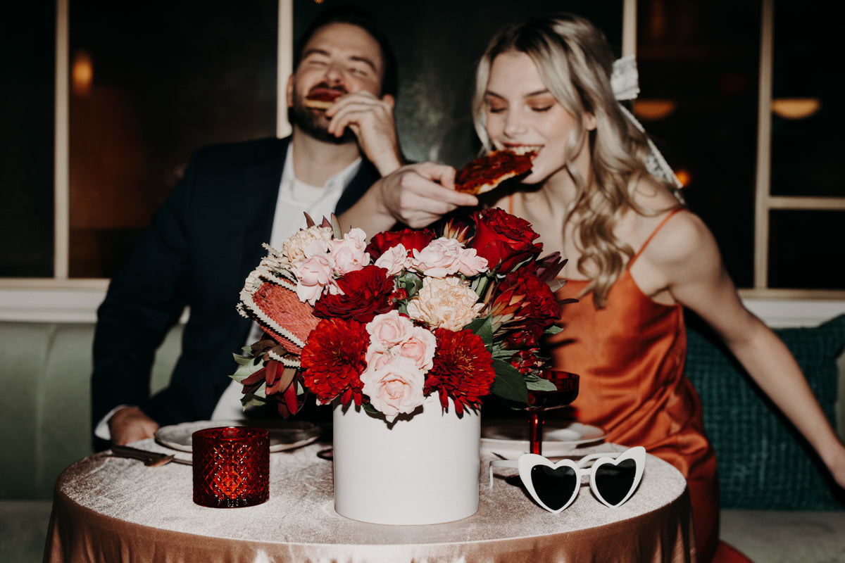 A newlywed couple playfully eats pizza in a retro-inspired Valentine’s shoot, with a dramatic red floral centerpiece setting the mood.