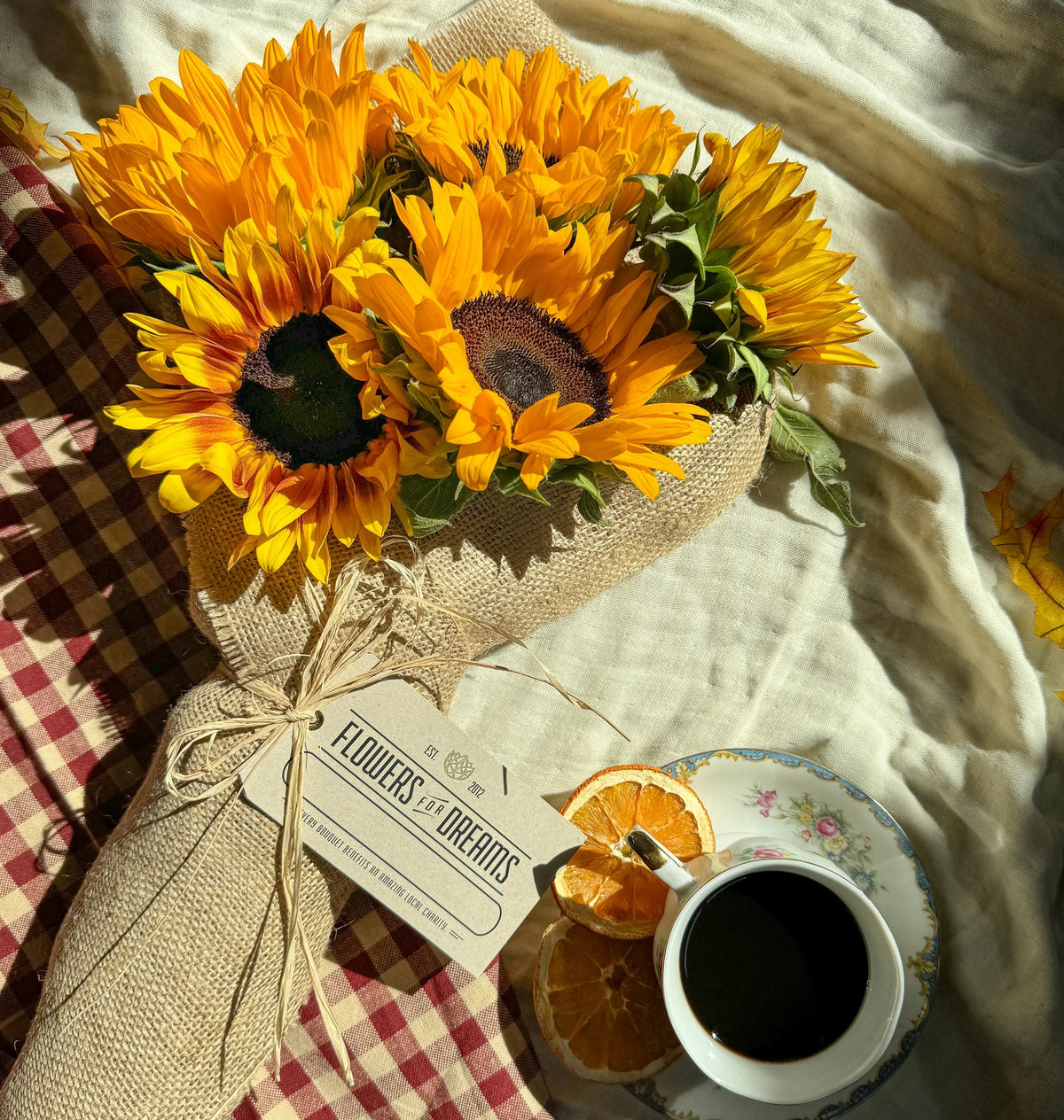 Sunflower bouquet from Flowers for Dreams wrapped in burlap with a coffee cup on checkered cloth.
