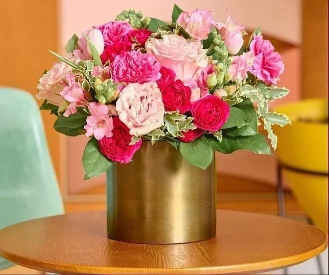 Brass vase filled with pink roses and carnations from Flowers for Dreams on a wooden table.