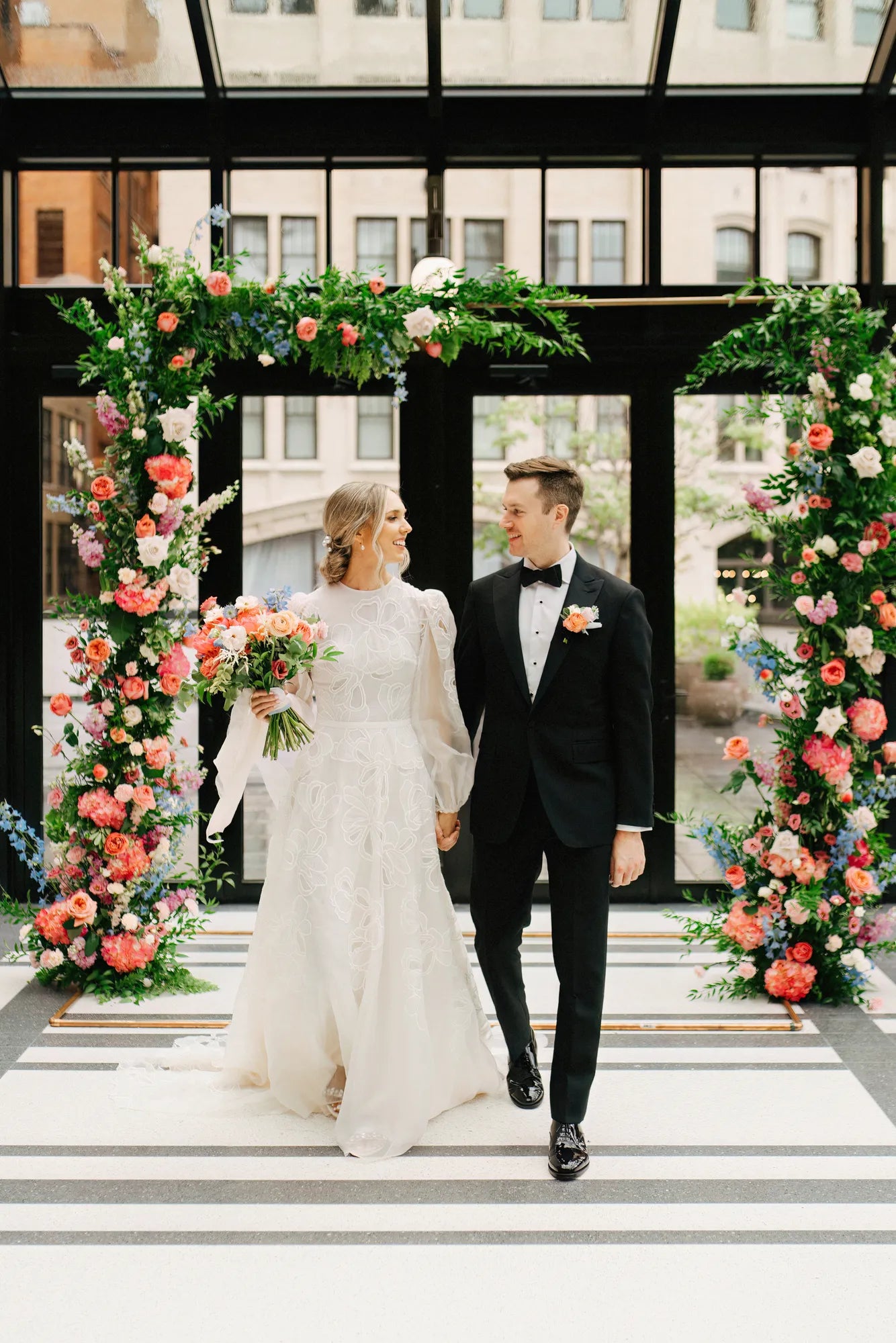 Bride and groom walk hand in hand beneath a lush floral arch with pink, coral, and blue blooms by Flowers for Dreams.