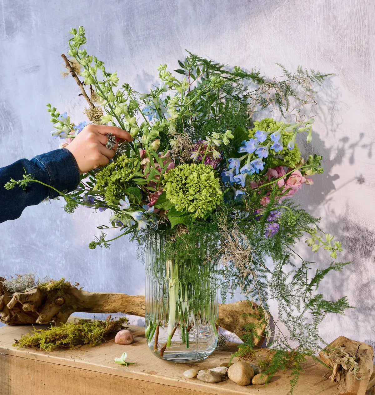A hand holding a colorful bouquet placed in a vase against a soft blue wall.