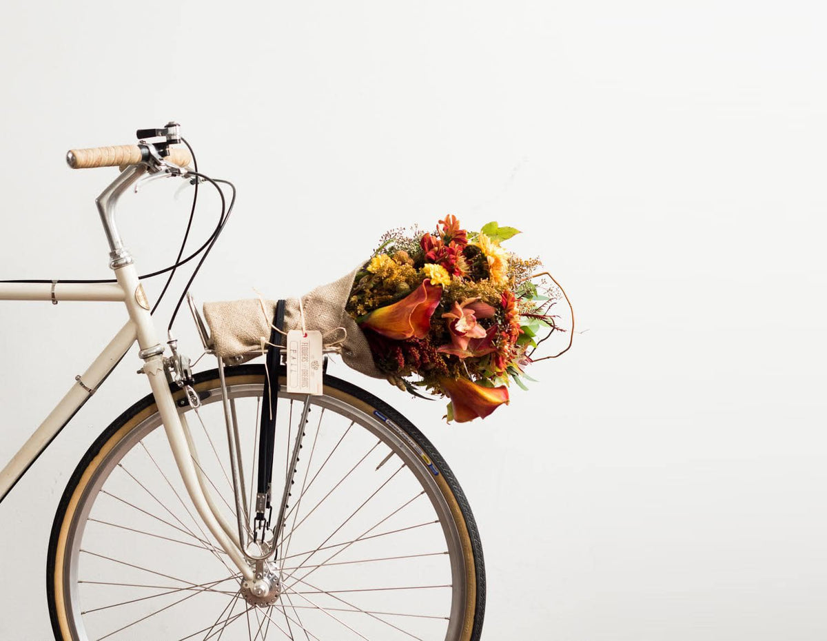 Beige vintage bicycle with a burlap-wrapped bouquet of autumn flowers secured to the rear rack, featuring warm orange and yellow tones.