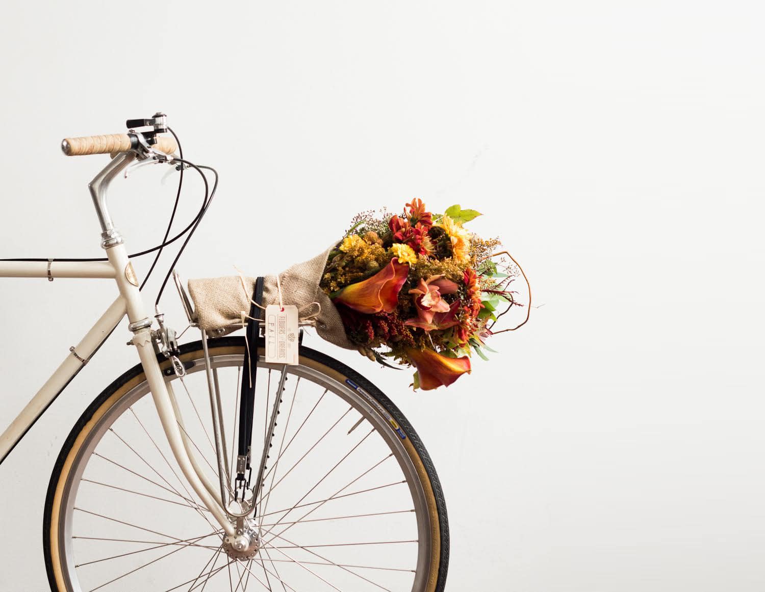 Beige vintage bicycle with a burlap-wrapped bouquet of autumn flowers secured to the rear rack, featuring warm orange and yellow tones.