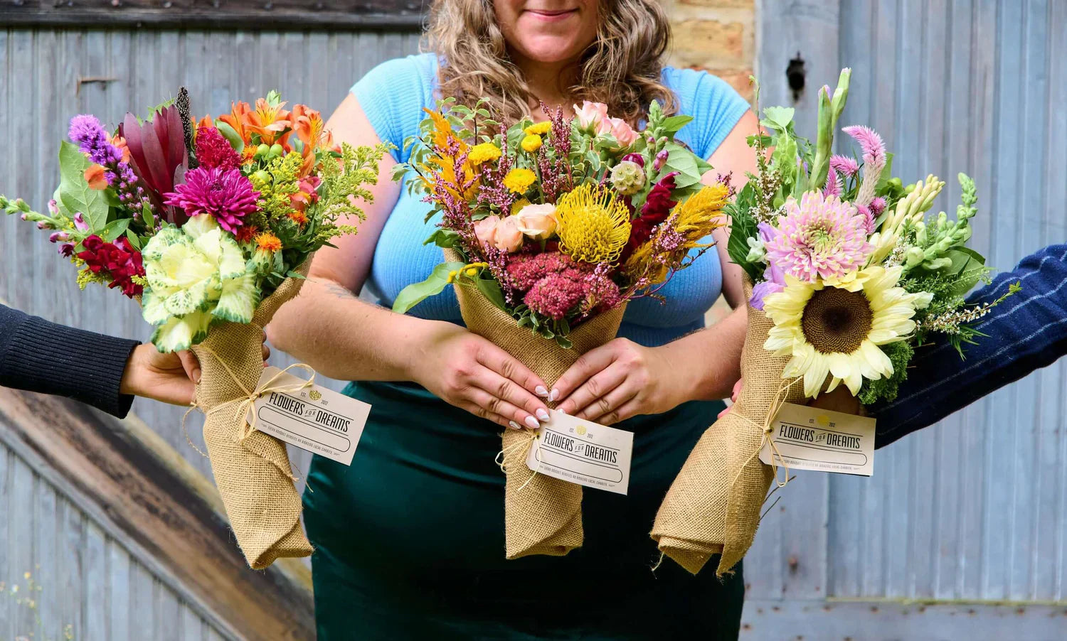 Three vibrant organic flower bouquets wrapped in burlap held by smiling hands from Flowers for Dreams.