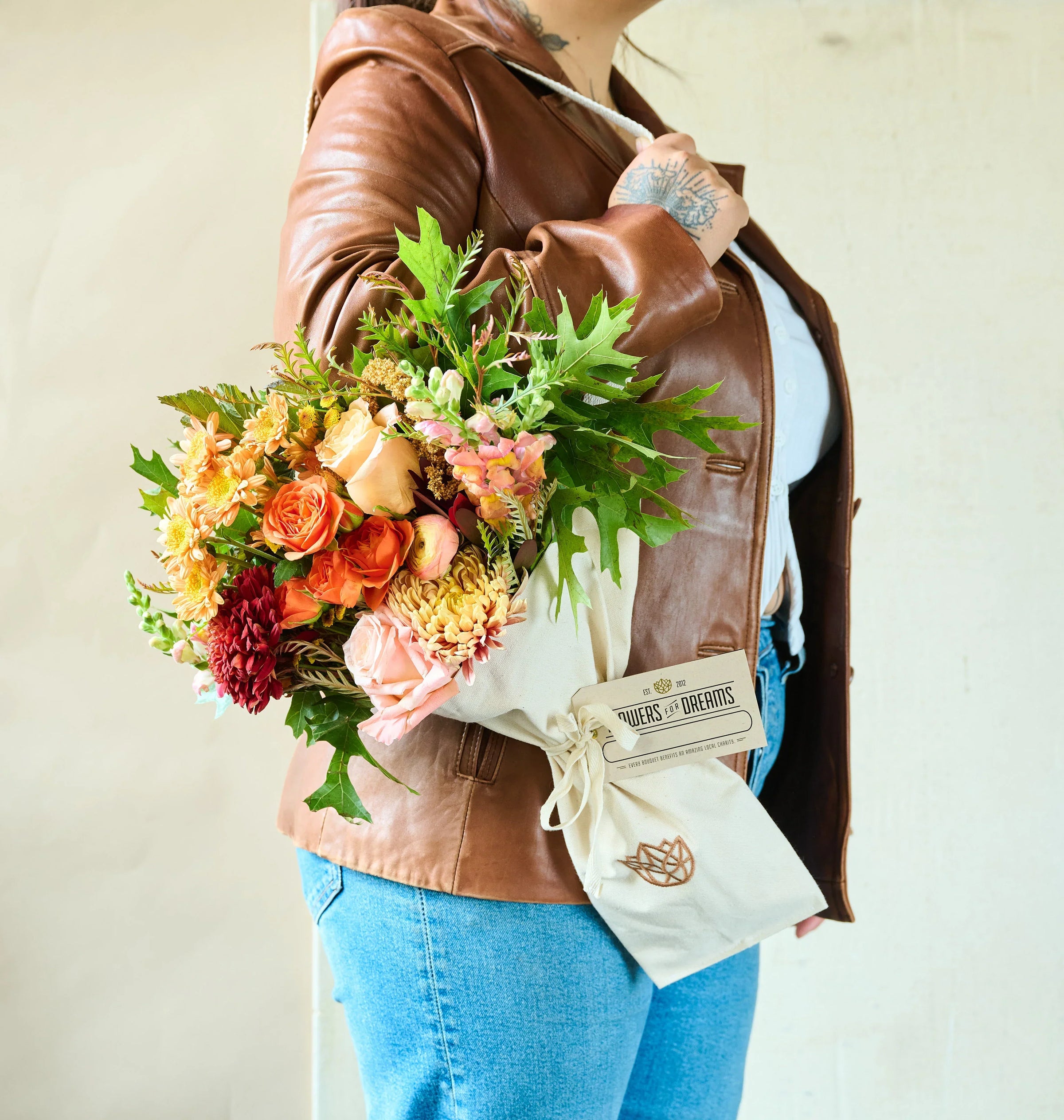 A person in a brown leather jacket holds a vibrant bouquet from Flower for Dreams of peach, orange, and red flowers. 