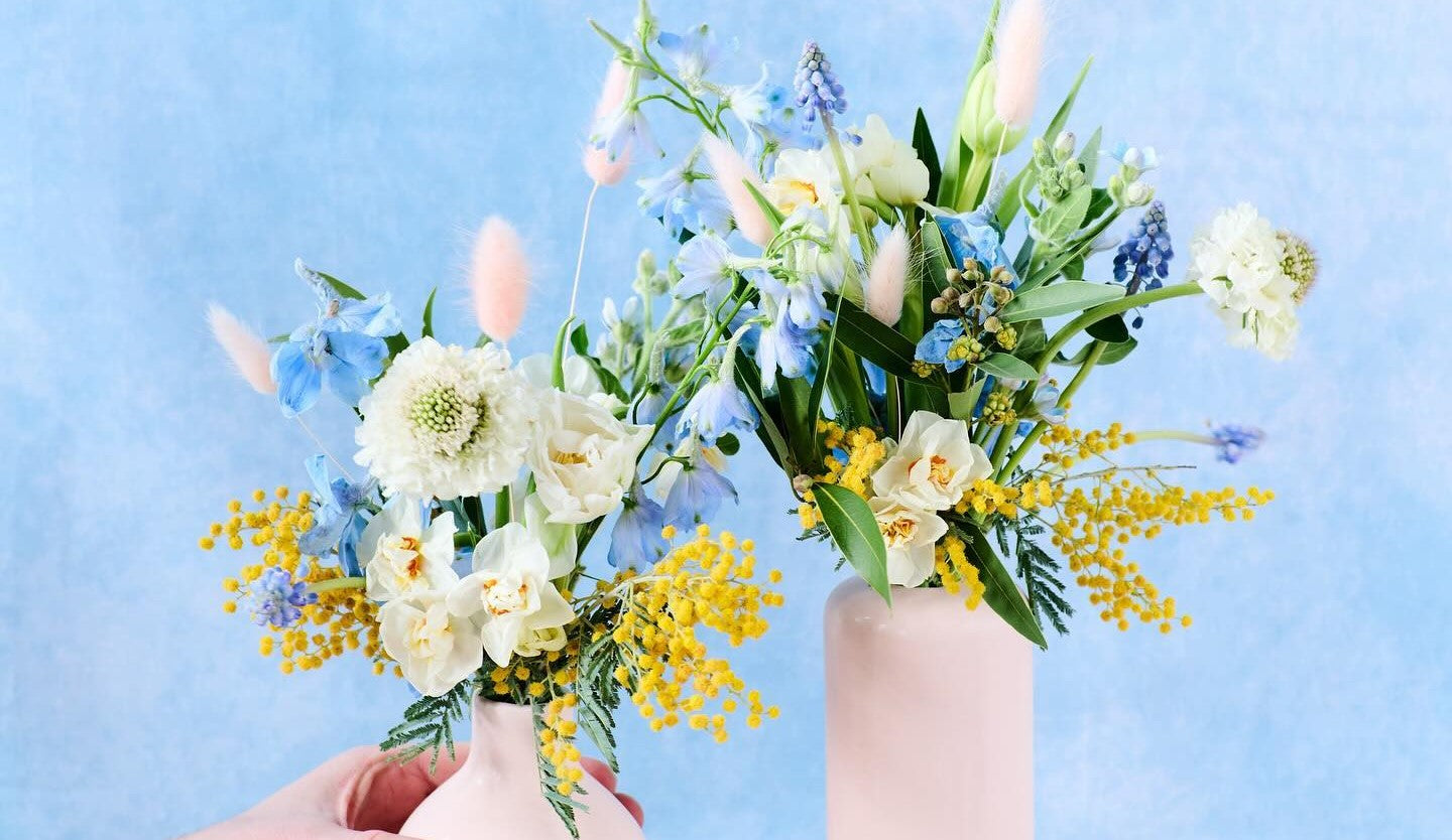 Pastel vases with spring blooms by Flowers for Dreams on a wooden tray, set against a soft blue backdrop.