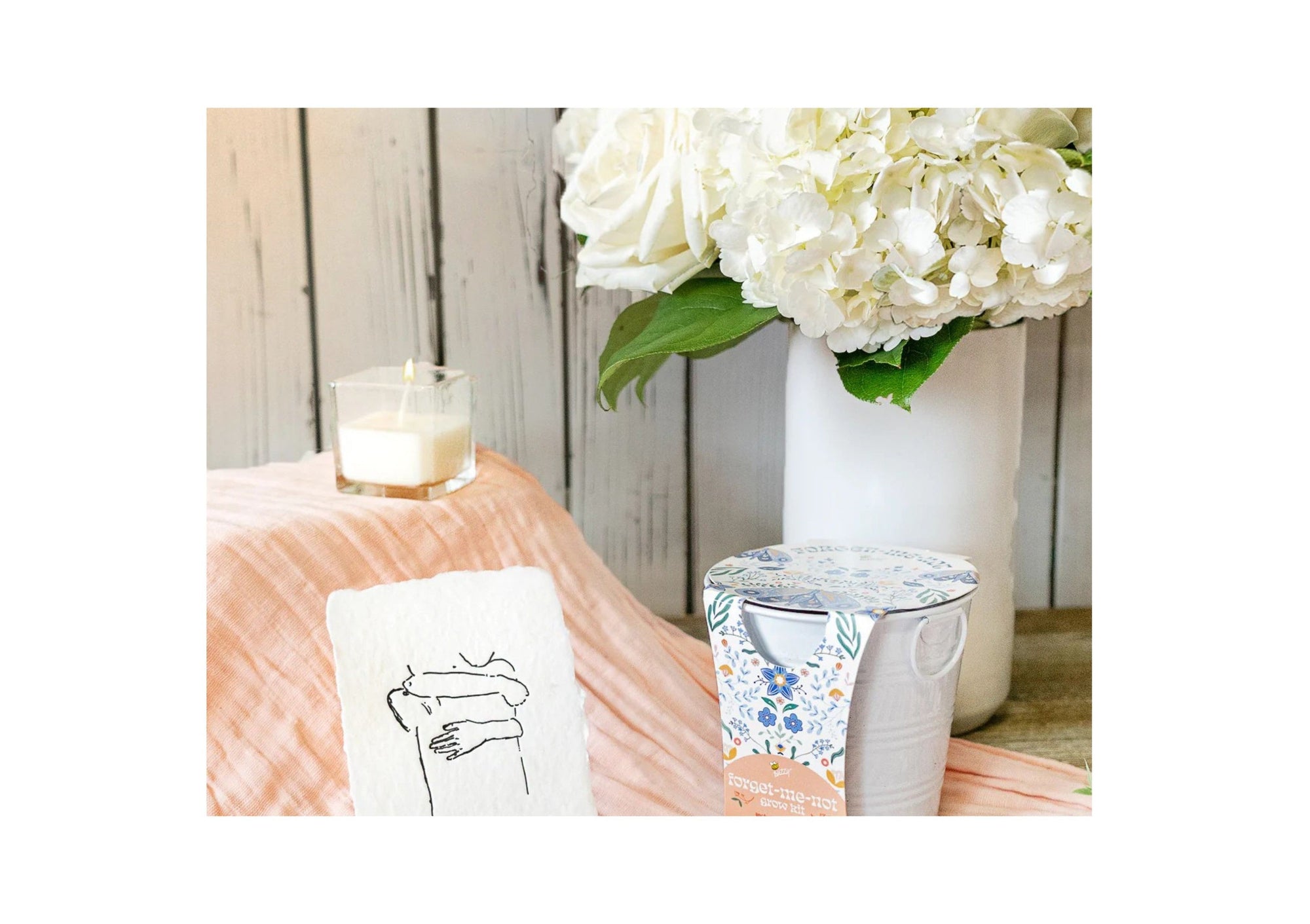 White hydrangea bouquet from Flowers for Dreams beside a candle, art card, and forget-me-not gift set.