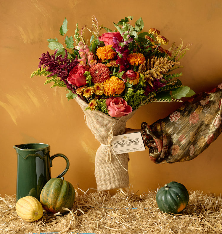 Flowers for Dreams Autumn bouquet of pink roses, orange marigolds, dahlias, chrysanthemums, amaranth, and snapdragons wrapped in burlap with gift tag, held against amber background with decorative gourds