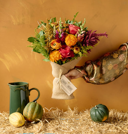 Autumn floral arrangement of pink roses, orange marigolds, dahlias, amaranth, and snapdragons in a white ceramic footed bowl on hay bale with decorative gourds, amber background