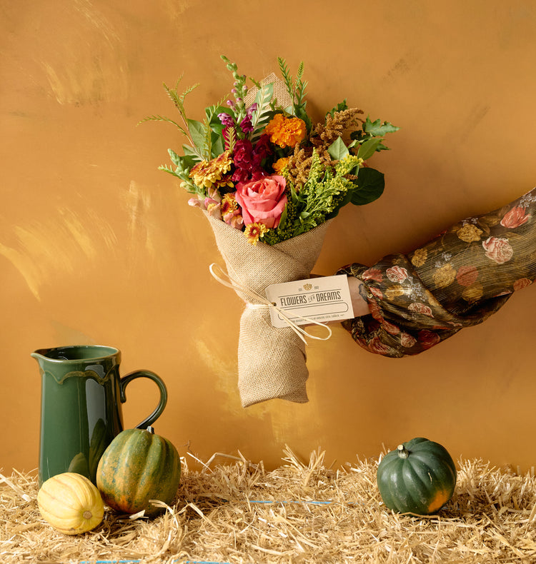 Autumn floral arrangement of pink roses, orange marigolds, dahlias, and amaranth in a white ceramic cylinder vase on hay bale with gourds, amber background
