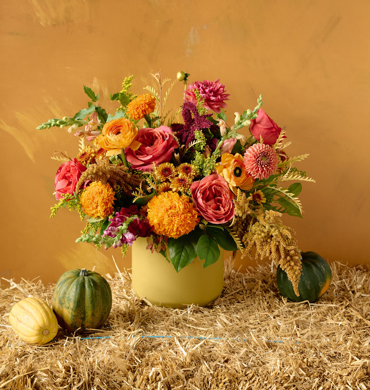 Autumn floral arrangement of pink roses, orange marigolds, chrysanthemums, and burgundy amaranth in a yellow ochre ceramic vase on hay bale with gourds, amber background