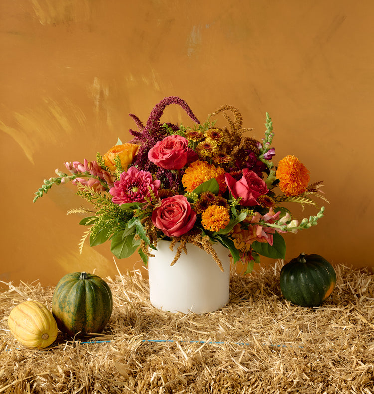 Autumn floral arrangement of pink roses, orange marigolds, chrysanthemums, burgundy amaranth, and snapdragons in a white ceramic cylinder vase on hay bale with gourds, amber background