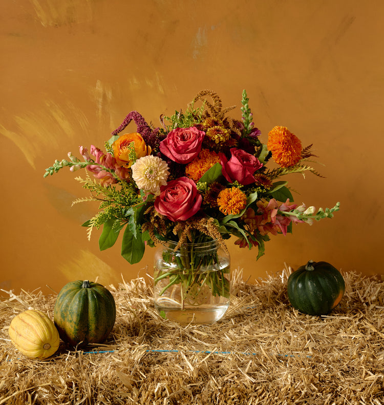 Autumn floral arrangement of pink roses, orange marigolds, ranunculus, white dahlias, chrysanthemums, and amaranth in a clear glass mason jar on hay bale with gourds, amber background