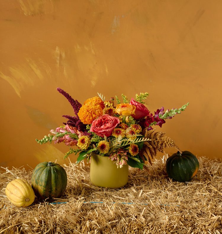 Autumn floral arrangement of pink roses, orange marigolds, dahlias, and burgundy amaranth in a bronze cylindrical vase on hay bale with gourds, amber background