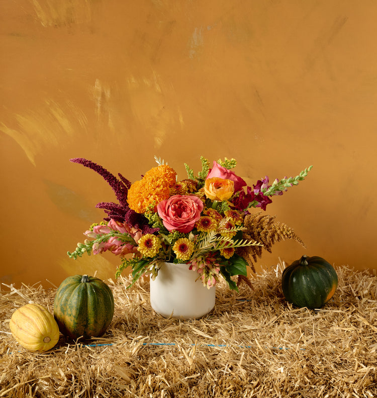 Autumn floral arrangement of pink roses, orange marigolds, chrysanthemums, snapdragons, and amaranth in a white ceramic low bowl on hay bale with decorative gourds, amber background