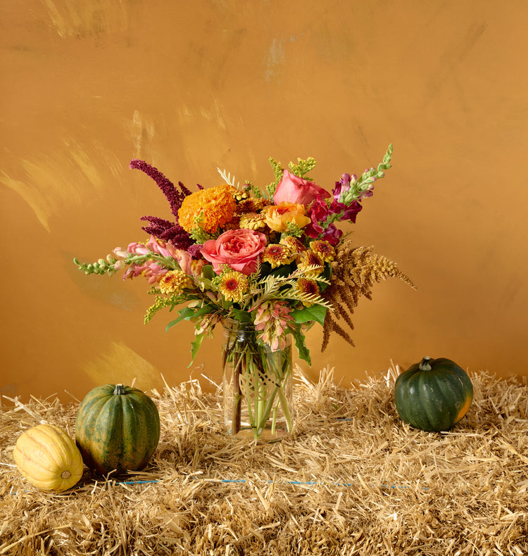 Autumn floral arrangement of pink roses, orange marigolds, ranunculus, chrysanthemums, and amaranth in a white ceramic bowl on hay bale with gourds, amber background