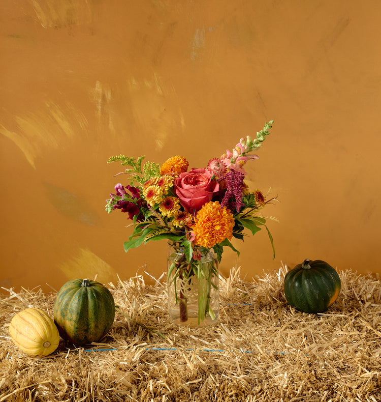 Autumn floral arrangement of pink roses, orange chrysanthemums, marigolds, and amaranth in a bronze cylindrical vase on hay bale with gourds, amber background