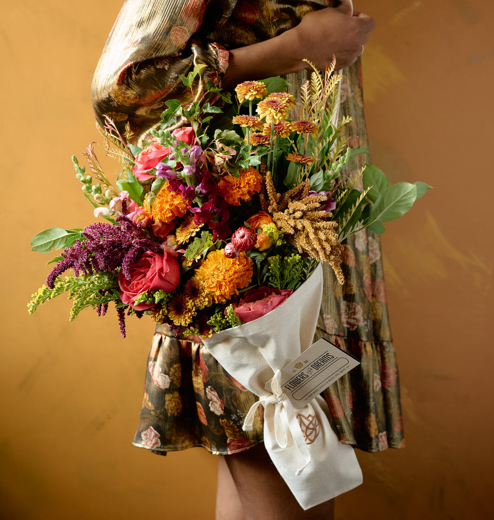 Person in floral dress holding large Flowers for Dreams Autumn bouquet of pink roses, orange marigolds, chrysanthemums, and mixed fall blooms wrapped in white ribbon, amber background