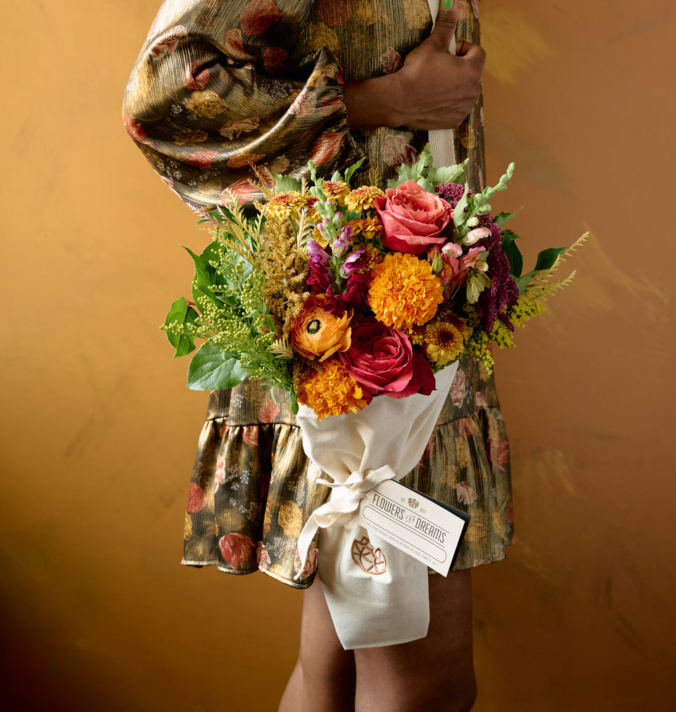Autumn floral arrangement of pink roses, orange marigolds, dahlias, and amaranth in a white tote bag vase on hay bale with gourds, amber background