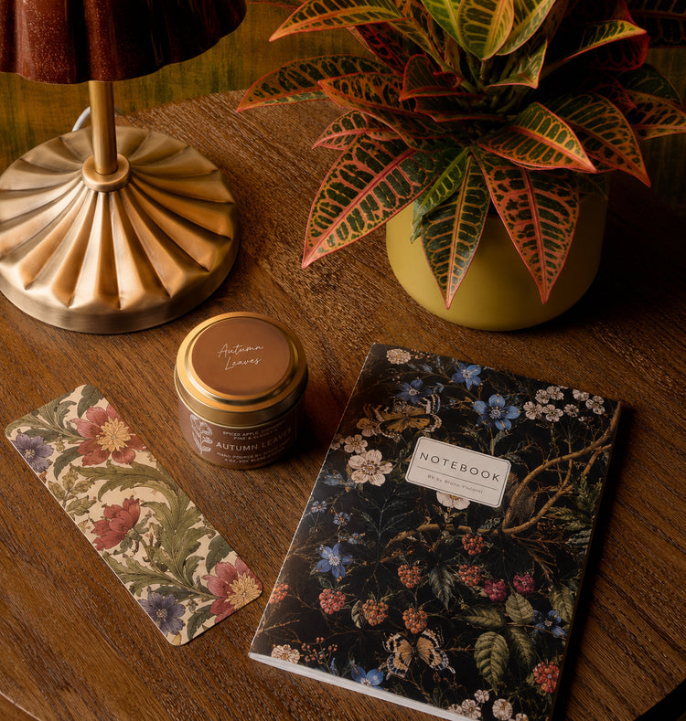 Cozy fall gift set flatlay featuring an Autumn Leaves soy candle tin, botanical floral notebook, bookmark, brass lamp, and croton plant on wood table