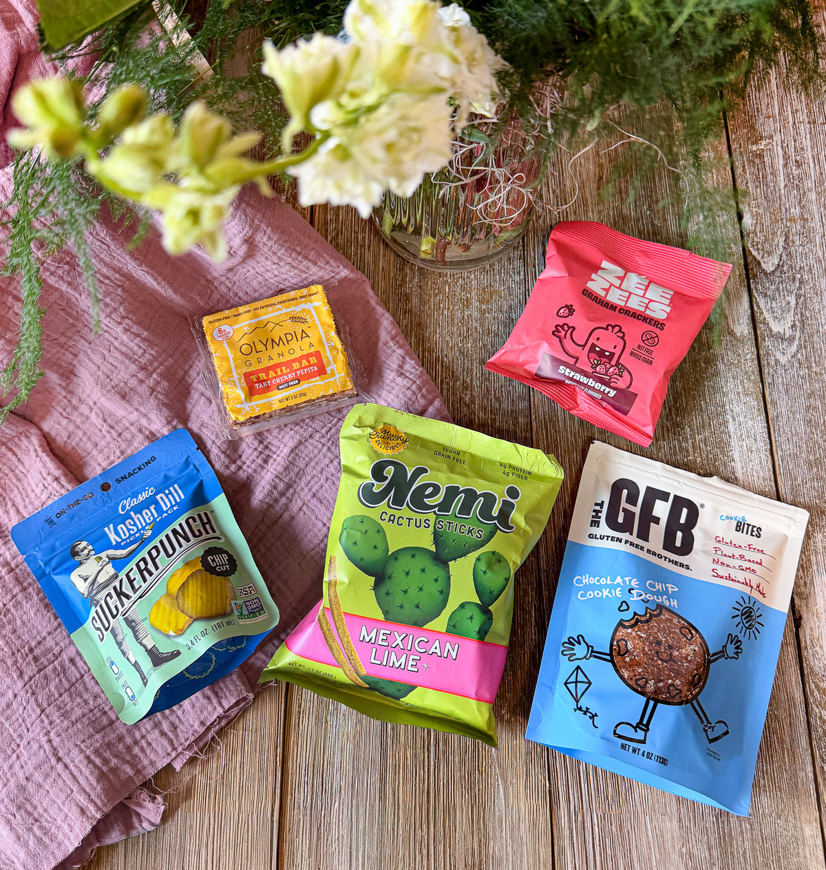 Five snack packets on a wooden surface with flowers and a pink cloth.