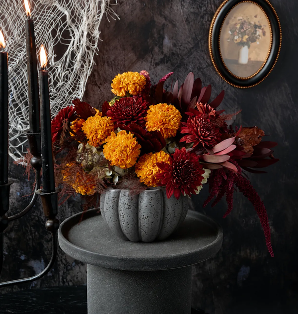 Haunt floral arrangement of orange marigolds, deep red chrysanthemums, and dark amaranth in a speckled grey pumpkin-shaped ceramic cauldron on dark Halloween backdrop with black candelabra