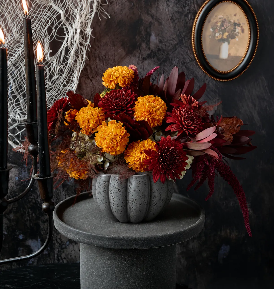 Haunt floral arrangement of orange marigolds, deep red chrysanthemums, and dark amaranth in a speckled grey pumpkin-shaped ceramic cauldron on dark Halloween backdrop with black candelabra